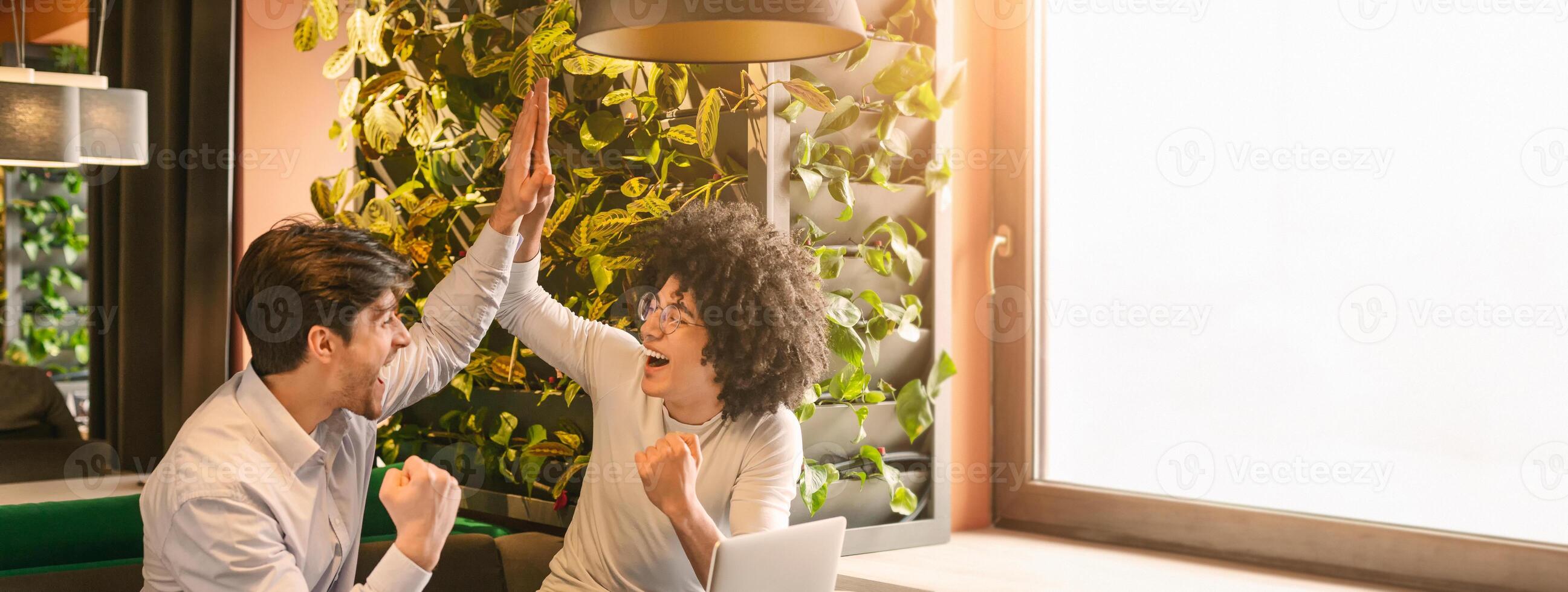 Two multiethnic colleagues express excitement and celebrate their success with high fives in a stylish workspace adorned with greenery, copy space photo