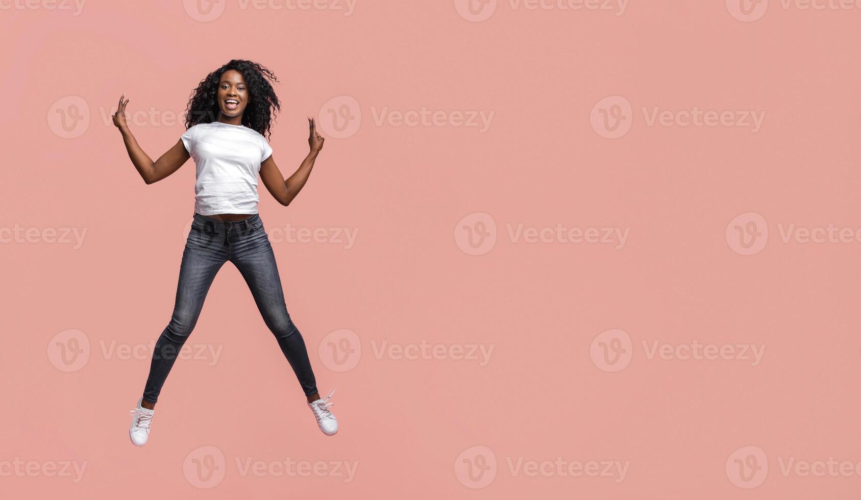 This image shows a young African American woman with curly hair jumping in front of a plain pink background. She is wearing a white t-shirt, dark blue jeans, and white sneakers. photo