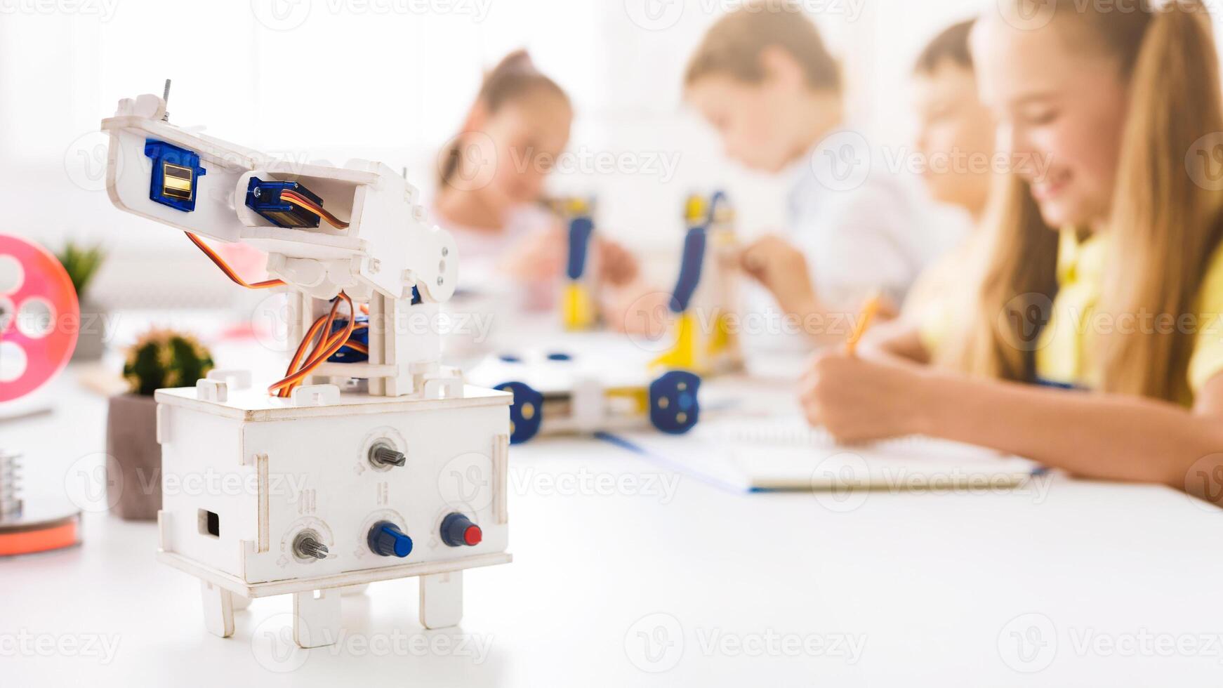 A close-up shot of a white robotic arm on a table in a classroom setting. The robot is in focus and there are blurred children in the background who are building or working on projects. photo