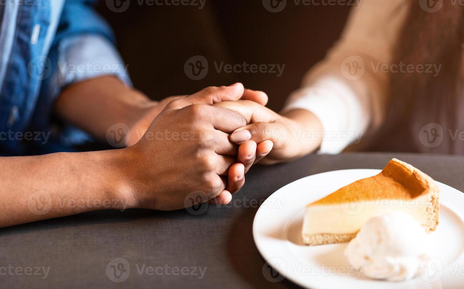 Two individuals are pictured holding hands at a table in a cozy cafe. A slice of cheesecake on a plate with a dollop of whipped cream is in front of them, suggesting a warm and intimate moment photo