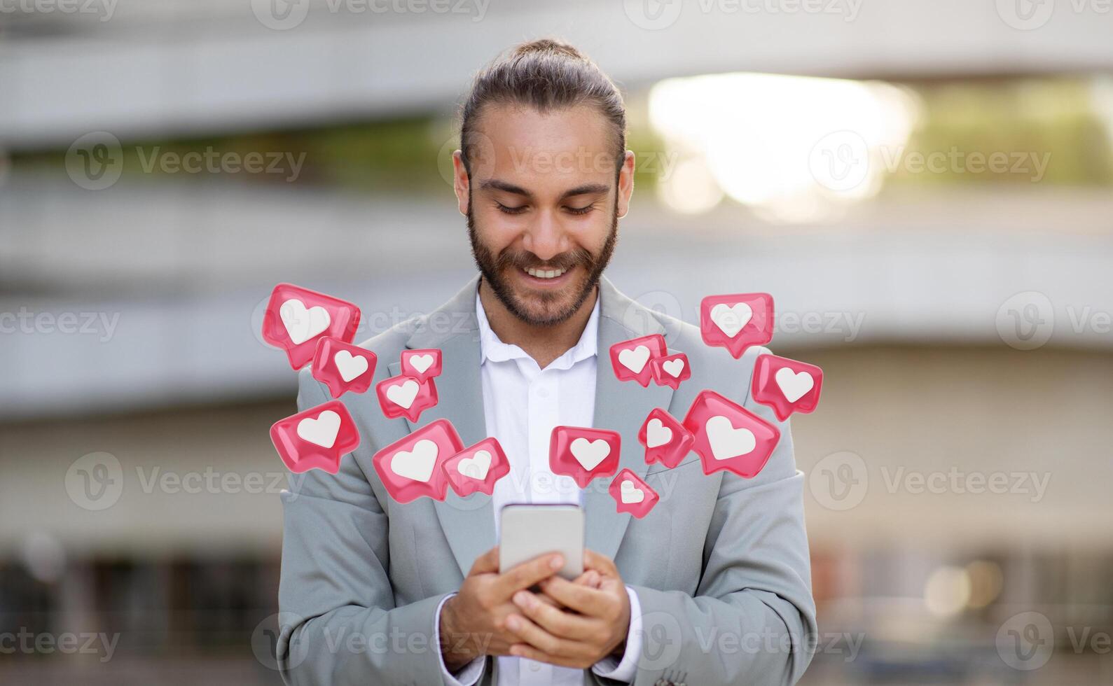A young man with a beard and a bun hairstyle is looking at his smartphone while smiling. He is wearing a gray suit jacket, and several digital hearts representing social media likes photo