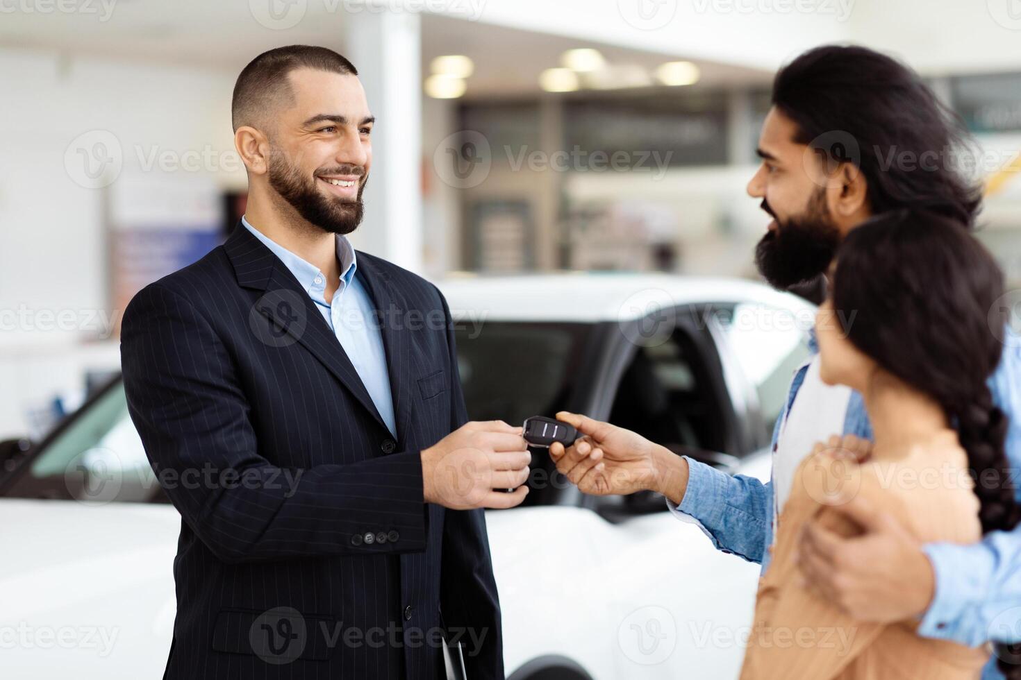 A well-dressed salesman is standing in a car showroom, presenting car keys to a cheerful Indian couple, symbolizing the finalization of a vehicle purchase photo