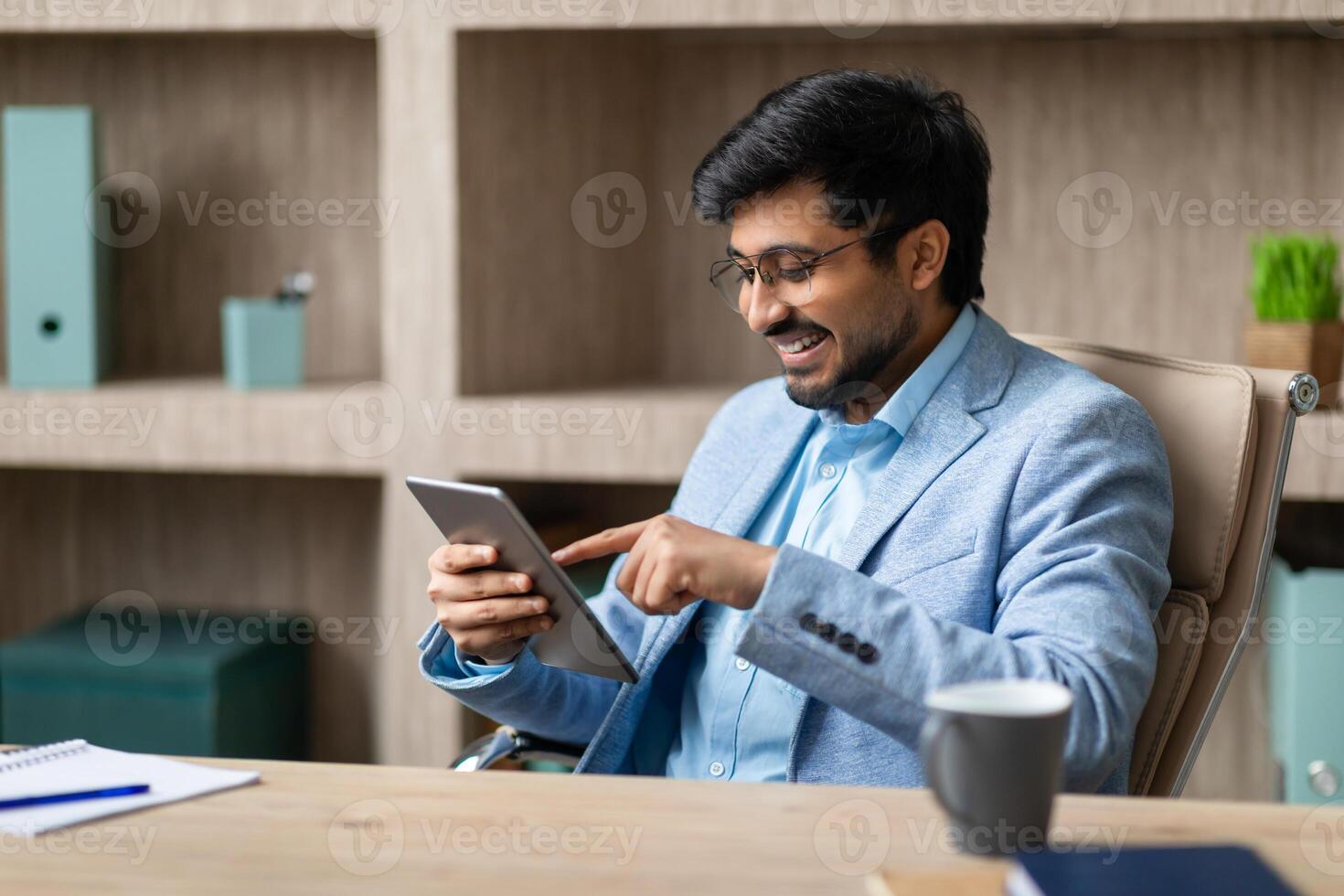 A smiling Indian businessman sits at a desk in his office, looking at a tablet and using his finger to navigate the screen. There is a cup of coffee on the desk and a notebook with a pen beside it. photo