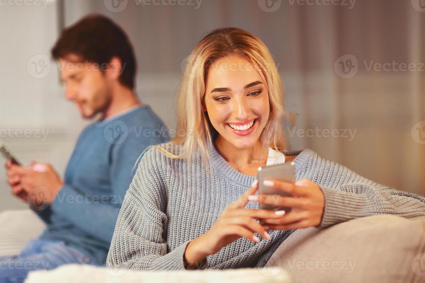 Cheating. Unfaithful Girlfriend Chatting On Phone With Lover Sitting Next To Boyfriend On Couch Indoor. Selective Focus photo