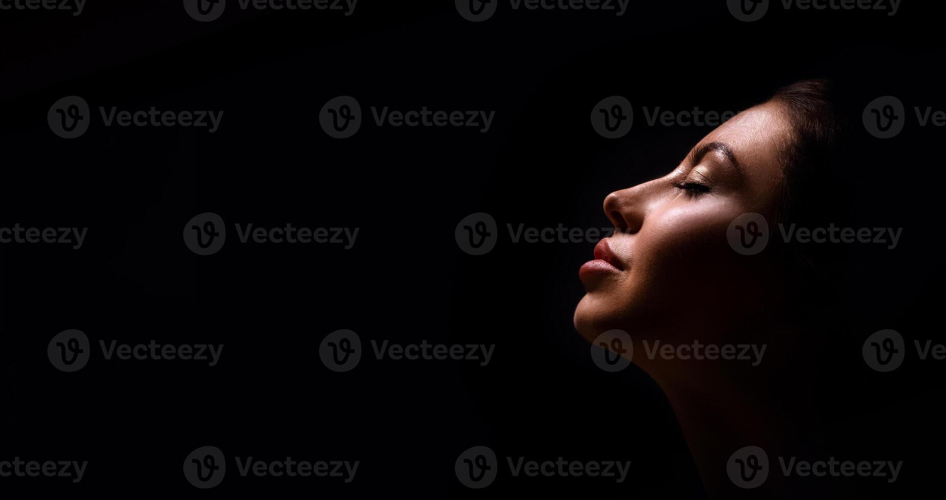 A close up shot of a womans face with eyes closed, lit from the side, with a dark background. She is turned slightly away from the camera. Her lips are closed and she appears to be relaxed. photo