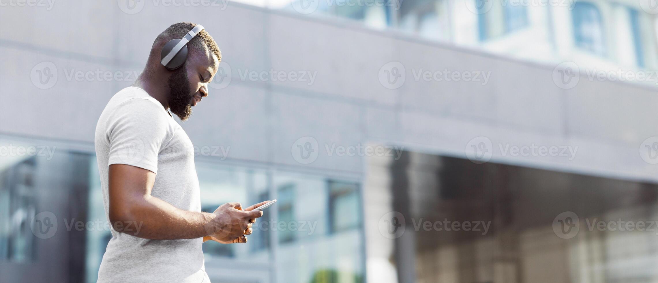 A man wearing headphones uses his phone while walking in an urban setting. photo