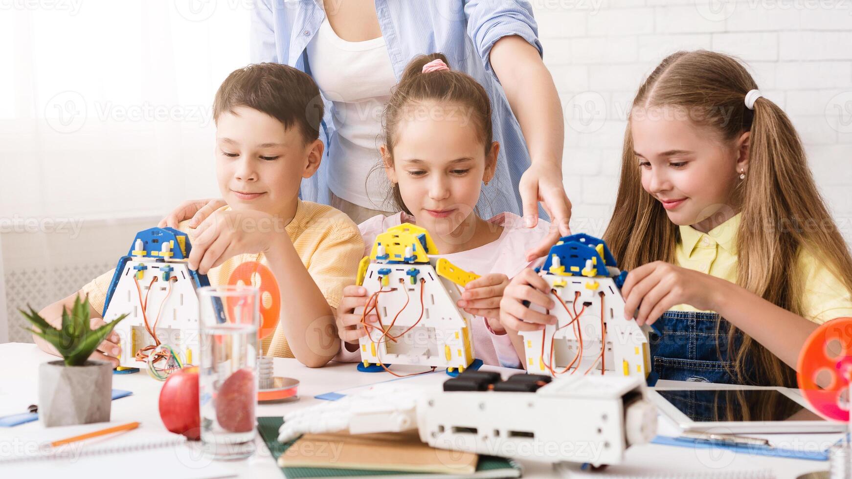 A group of young students are working together to build robots in a classroom setting. They are being guided by a teacher, who is standing behind them and observing their progress. photo