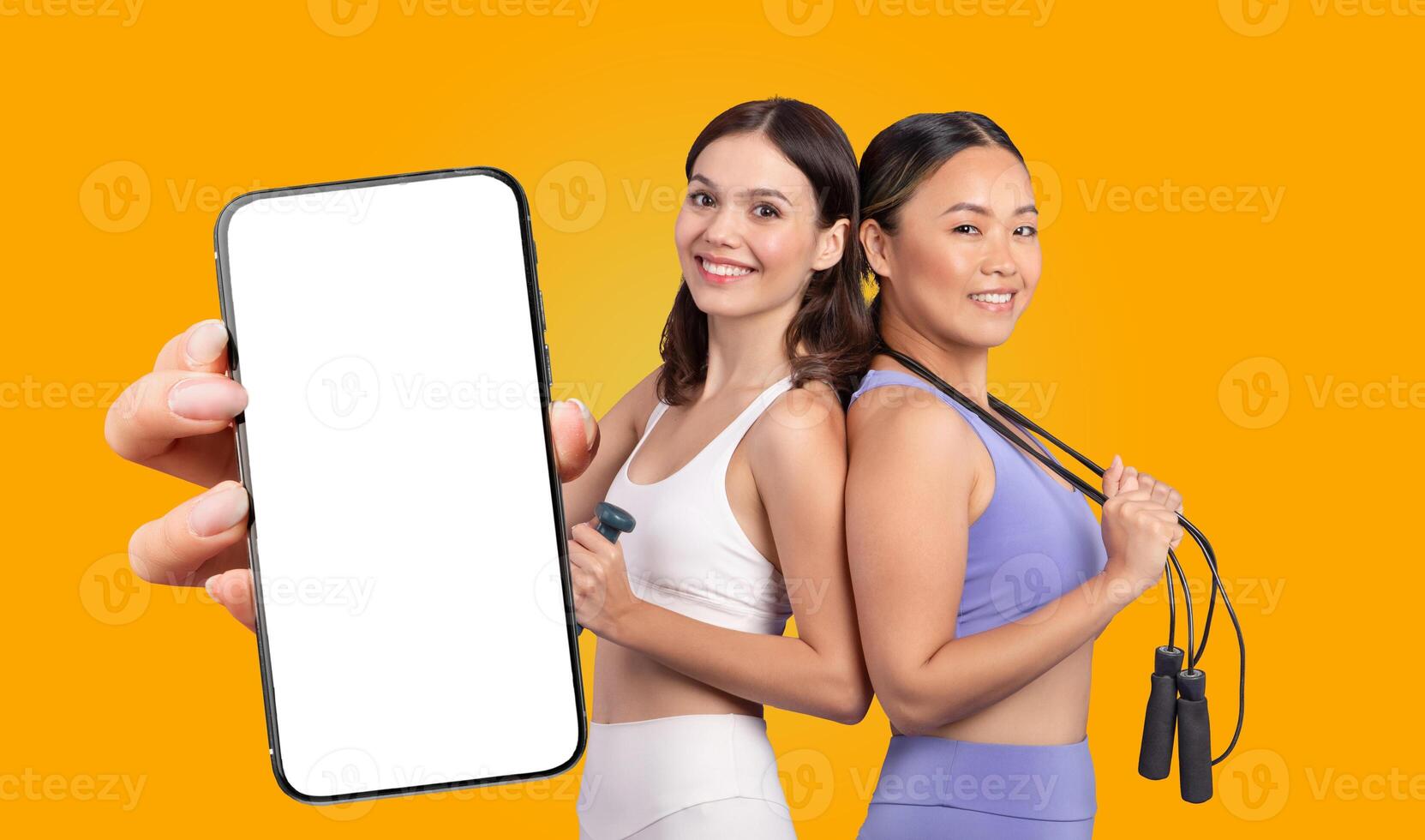Two multiethnic women wearing athletic wear are smiling and standing side by side. The woman on the left is holding a smartphone with a blank screen while woman on the right is holding a jump rope. photo