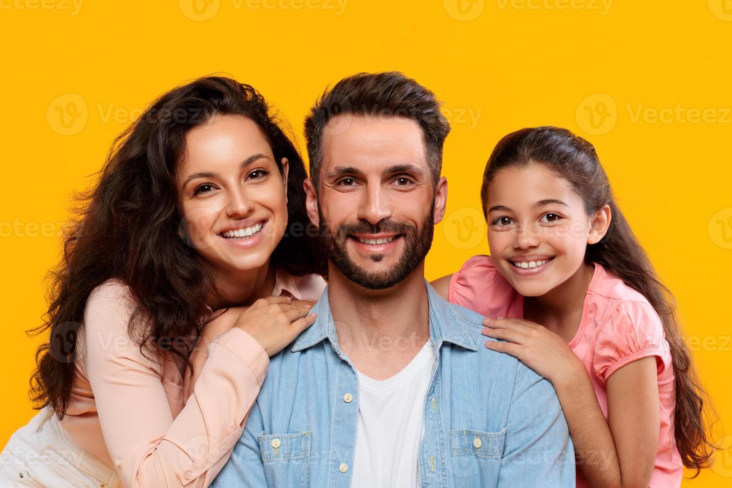 A smiling family of three poses for a portrait against a bright yellow backdrop. Father, mother and daughter enjoying time together photo