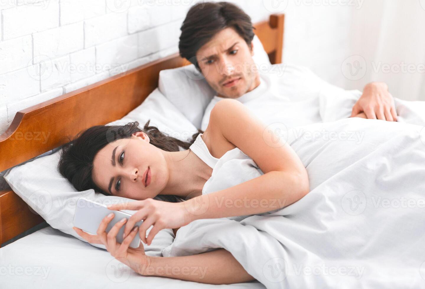 A woman is lying in bed, looking at her phone while a man, who is also in bed, watches her suspiciously. This image can be used to illustrate infidelity, mistrust, or relationship problems. photo