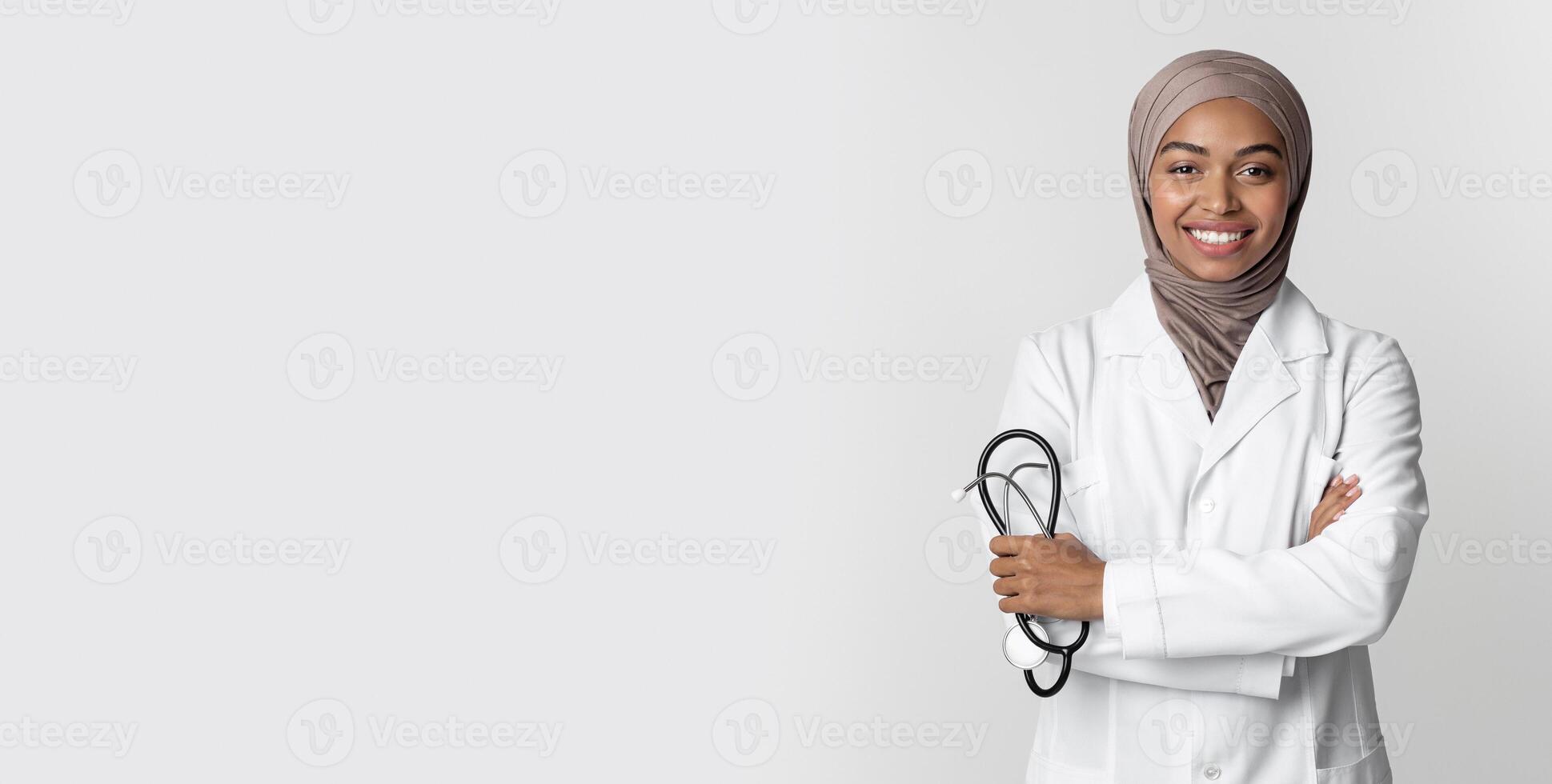 Portrait Of Young Afro Muslim Female Doctor In Hijab Wearing White Coat And Holding Stethoscope, Posing With Folded Arms Over Light Background photo