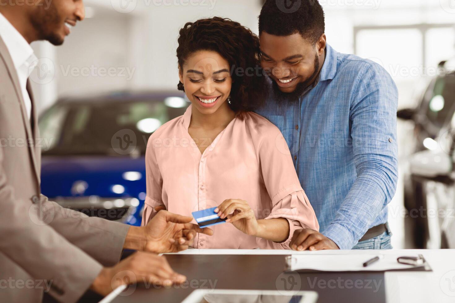 Buying Car Concept. Smiling Black Spouses Giving Credit Card To Automobile Seller In Auto Selling Showroom. Selective Focus photo