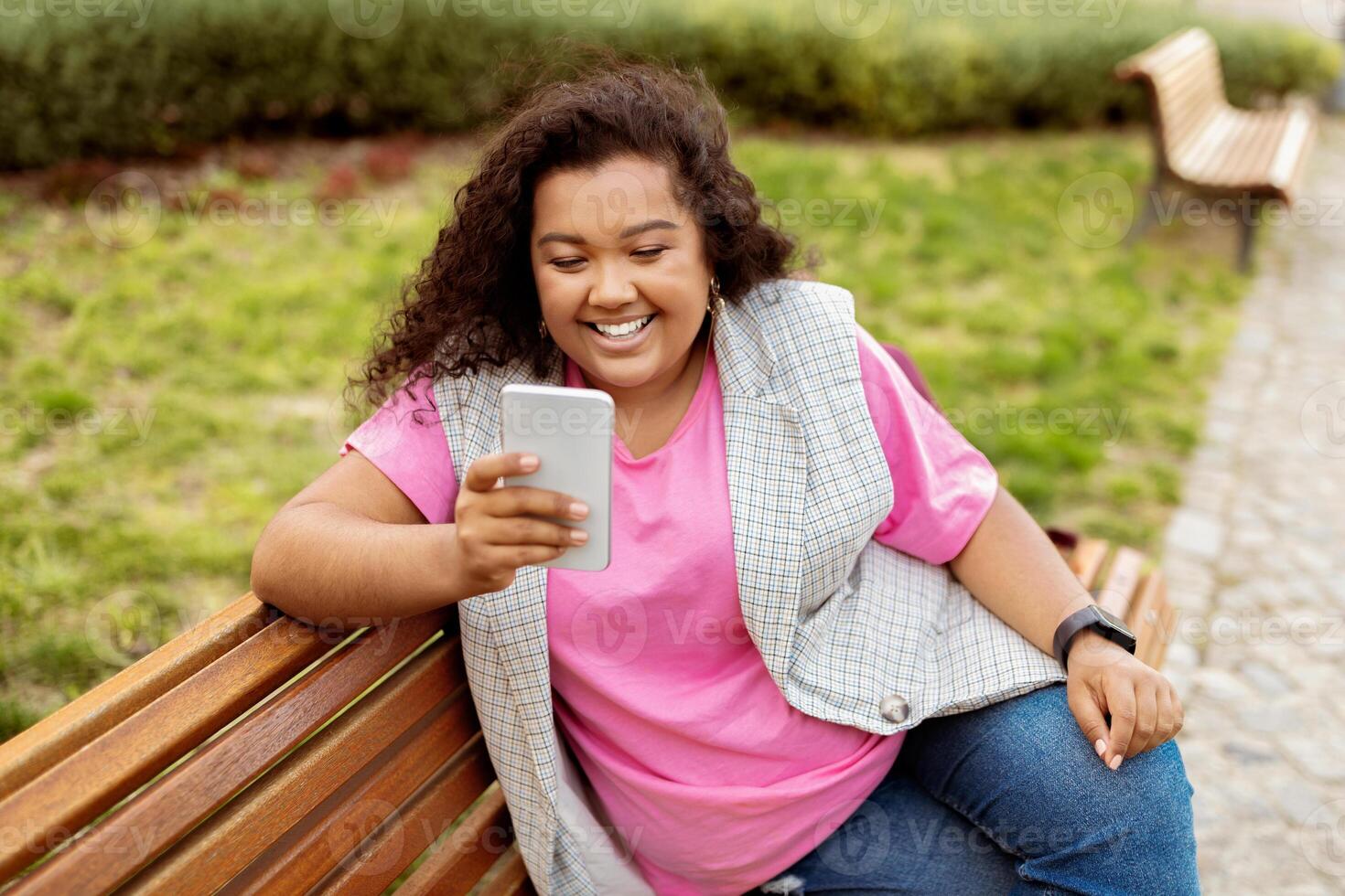 A young woman with curly hair sits on a bench in a green park, smiling as she looks at her smartphone, enjoying a relaxed moment outdoors on a sunny day. photo