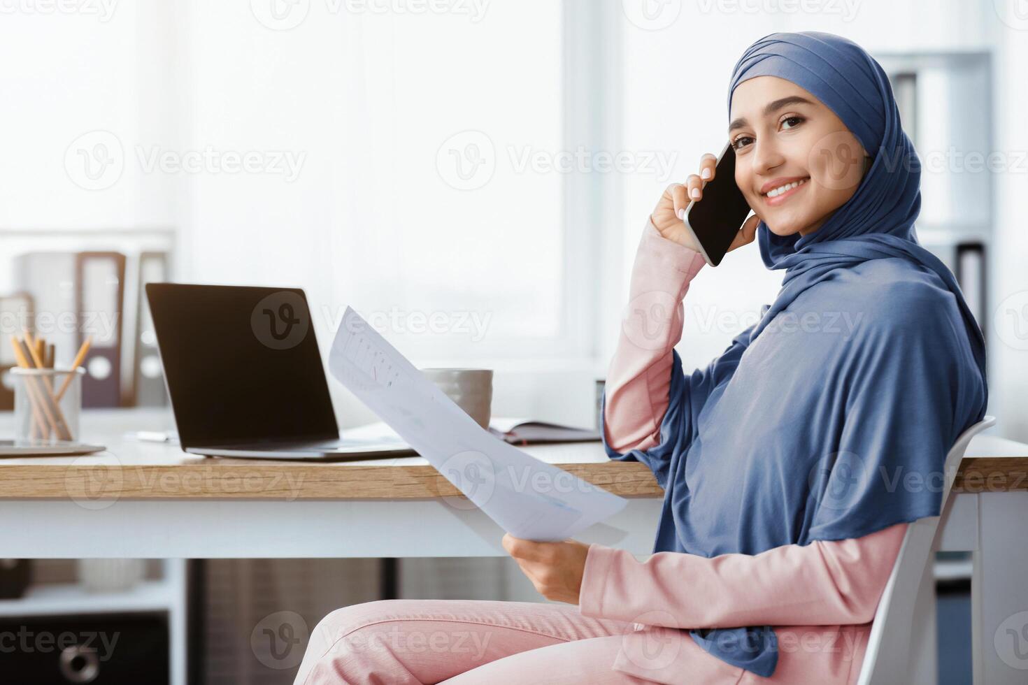 Smiling saudi businesswoman in hijab working with documents and talking on cellphone at workplace in office, side view with empy space photo