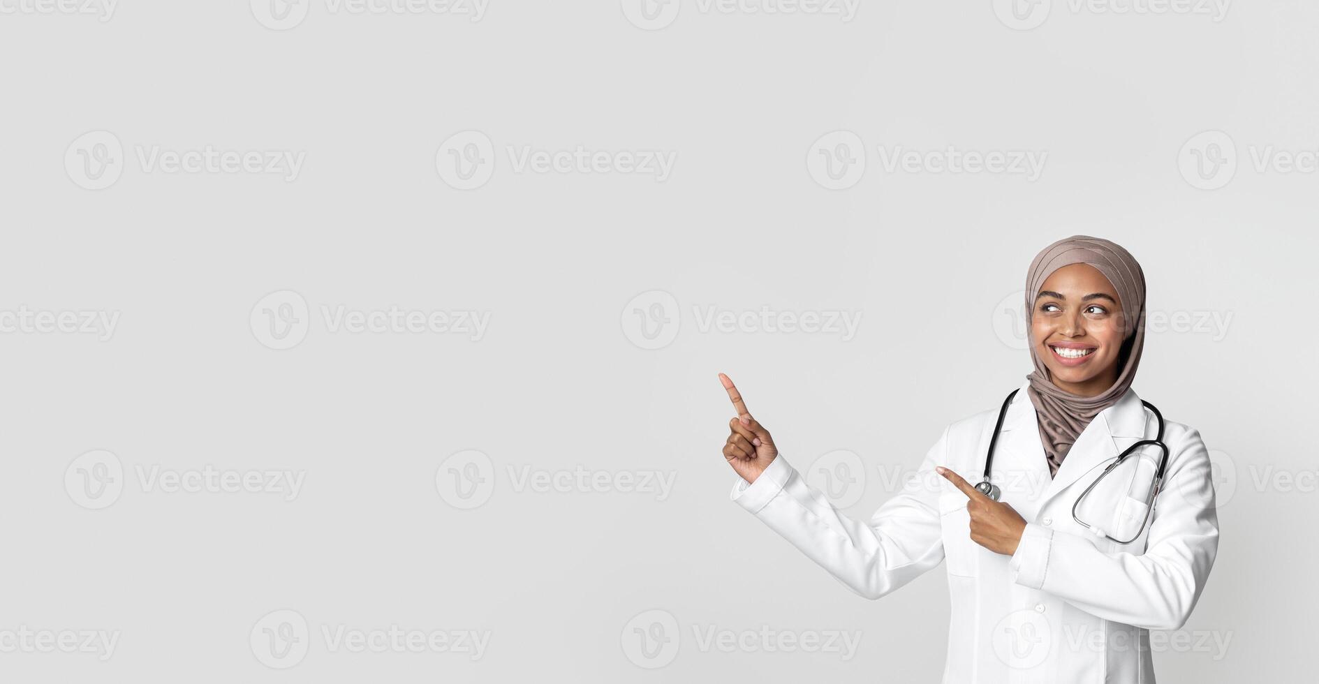 Medical Ad. Young black female therapist in hijab and white coat with stethoscope pointing at copy space over light background with her index fingers photo