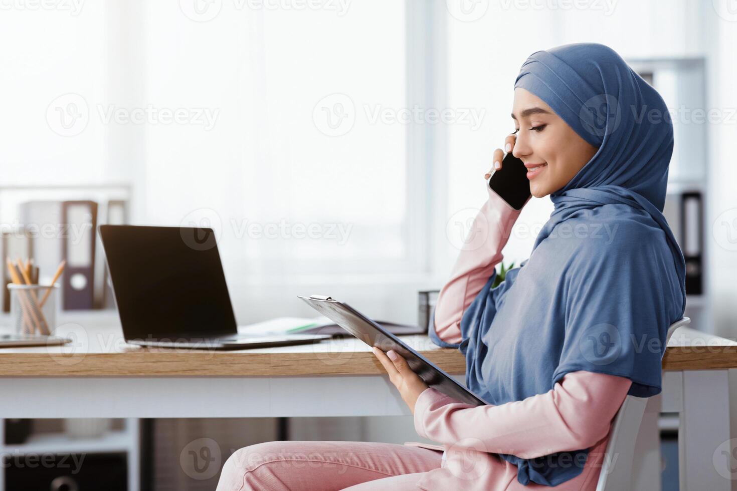 Muslim business lady in hijab holding clipboard, checking documentation and talking on mobile phone with partner while sitting at workplace in office photo