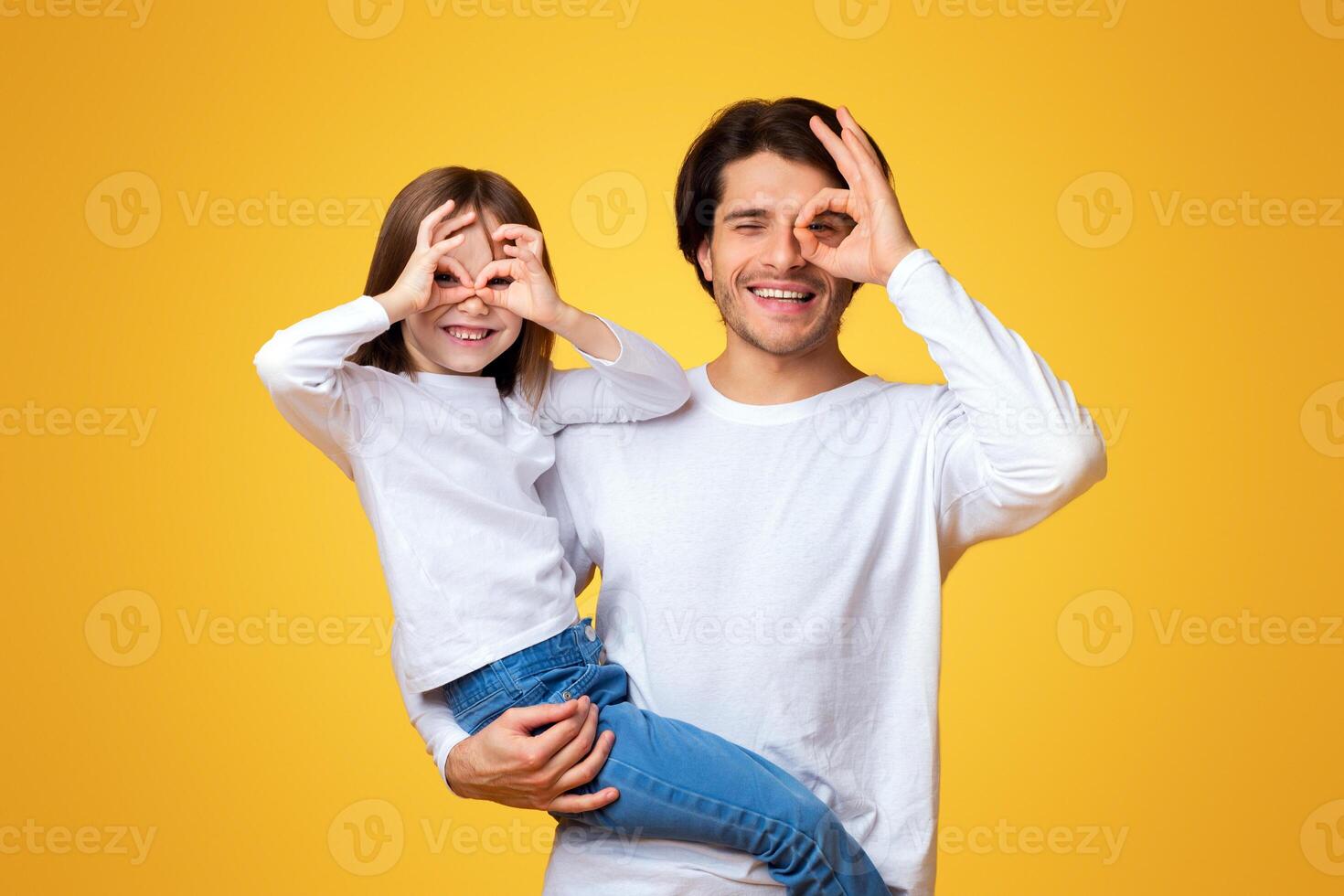 Funny little girl and daddy fooling, having fun over yellow background, imitating binoculars photo