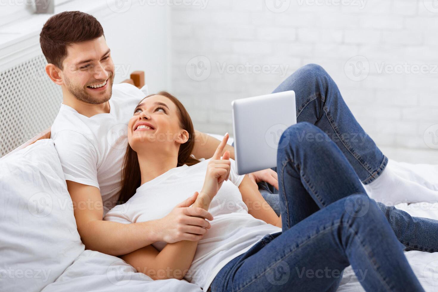 Cheerful Spouses Using Tablet Computer Lying In Bed At Home Relaxing On Weekend Morning. Selective Focus photo