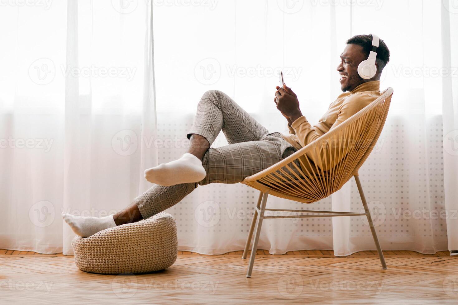 Weekend. Cheerful Afro Man In Wireless Earphones Using Cellphone Listening To Music Sitting On Modern Chair Against Window At Home photo