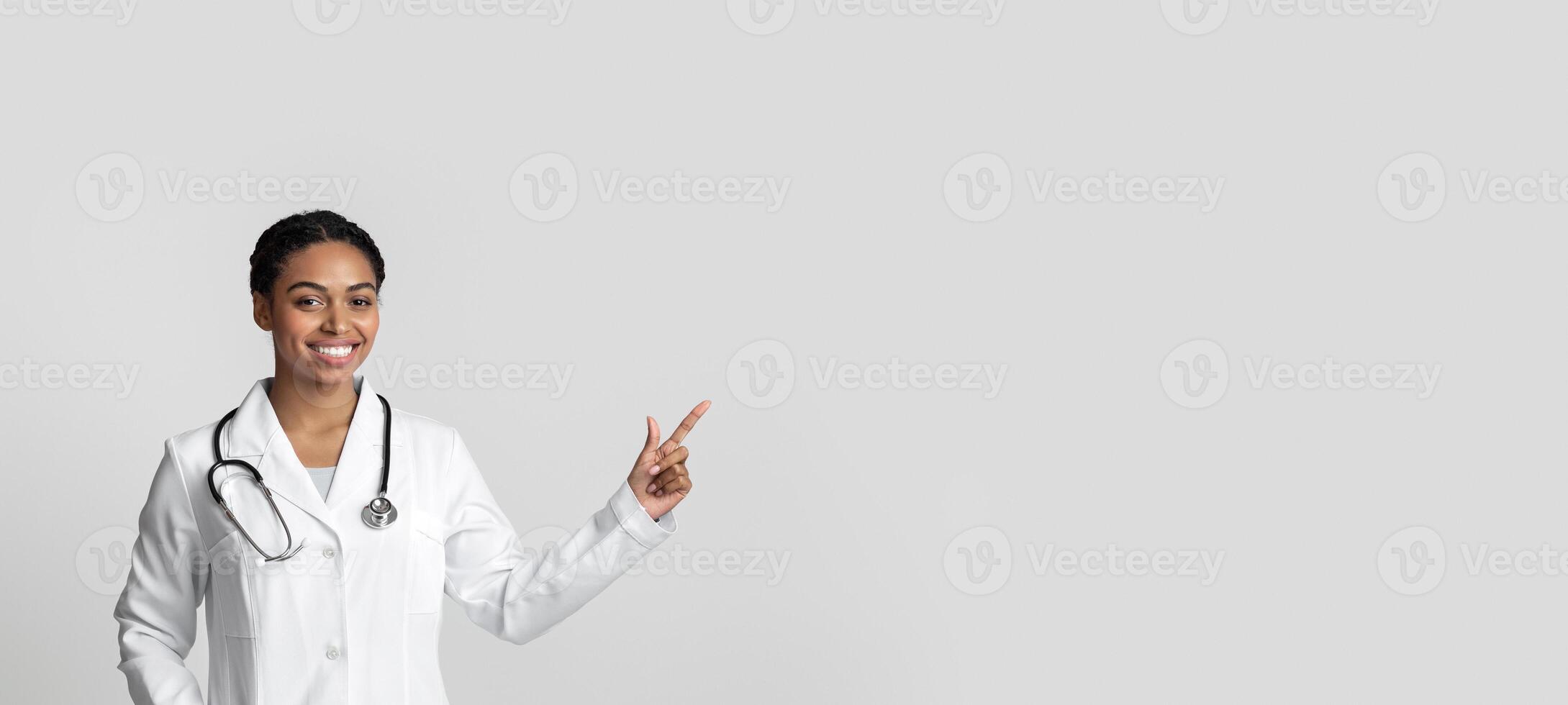 Young black female doctor pointing at copy space on light background, wearing white coat and stethoscope and smiling at camera photo