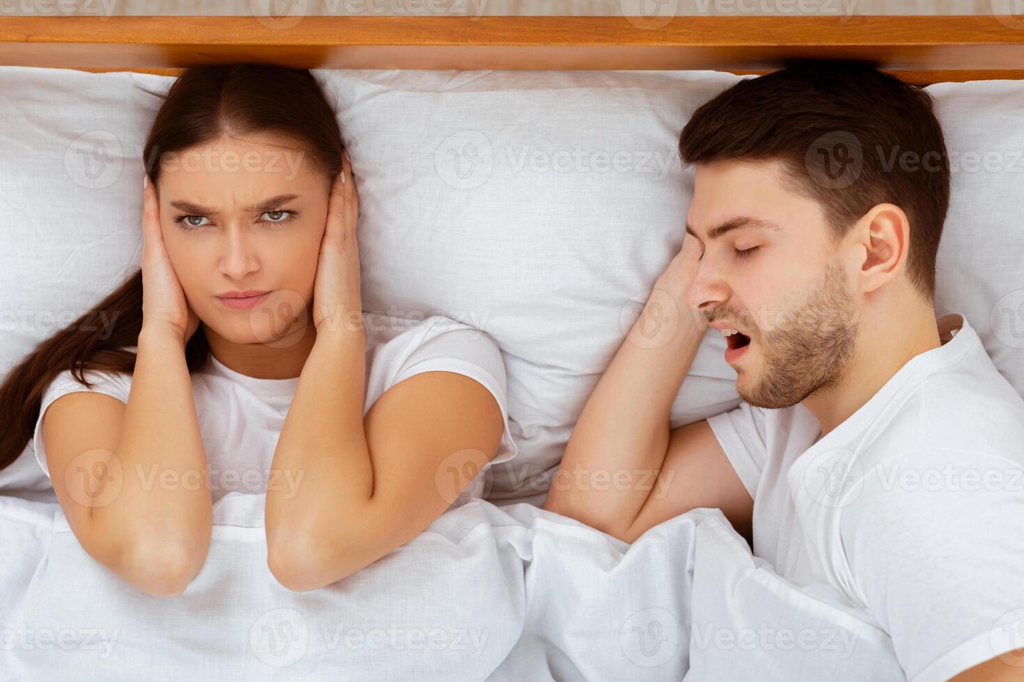 Snorting Problem. Angry Wife Covering Ears Trying To Sleep Near Snoring Husband In Bed At Home. Selective Focus, Top View photo