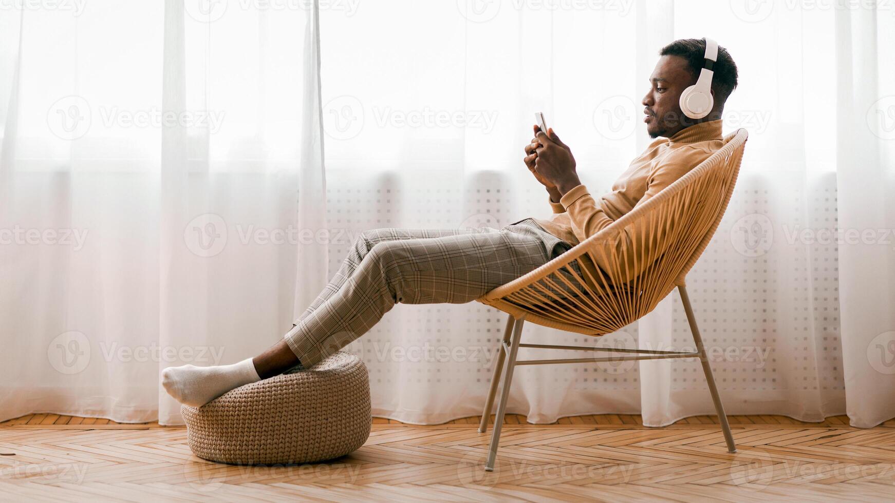Weekend. Relaxed Black Man In Wireless Headset Using Mobile Phone Listening To Audiobook Sitting On Modern Chair Against Window Indoor photo