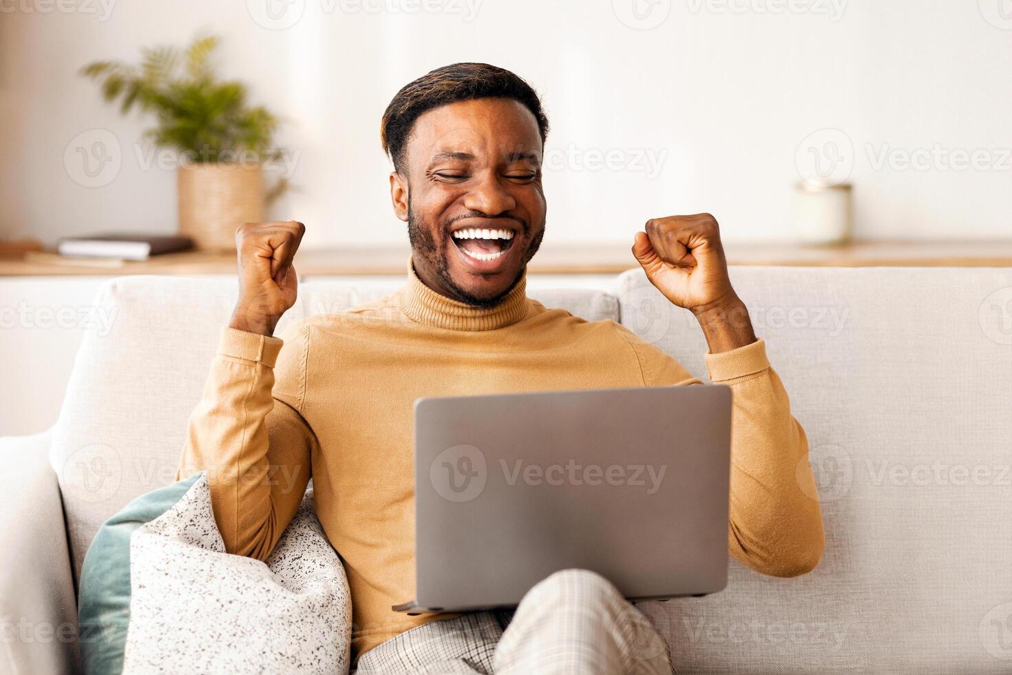 Business Success. Excited Black Man Using Laptop Gesturing Yes Celebrating Great News Sitting On Couch Indoor. Copy Space photo