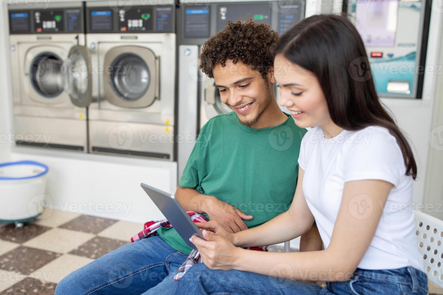 Two young adults sit together in a laundromat, smiling as they look at a tablet. The laundry machines are in the background, indicating a relaxed atmosphere while they wait. photo