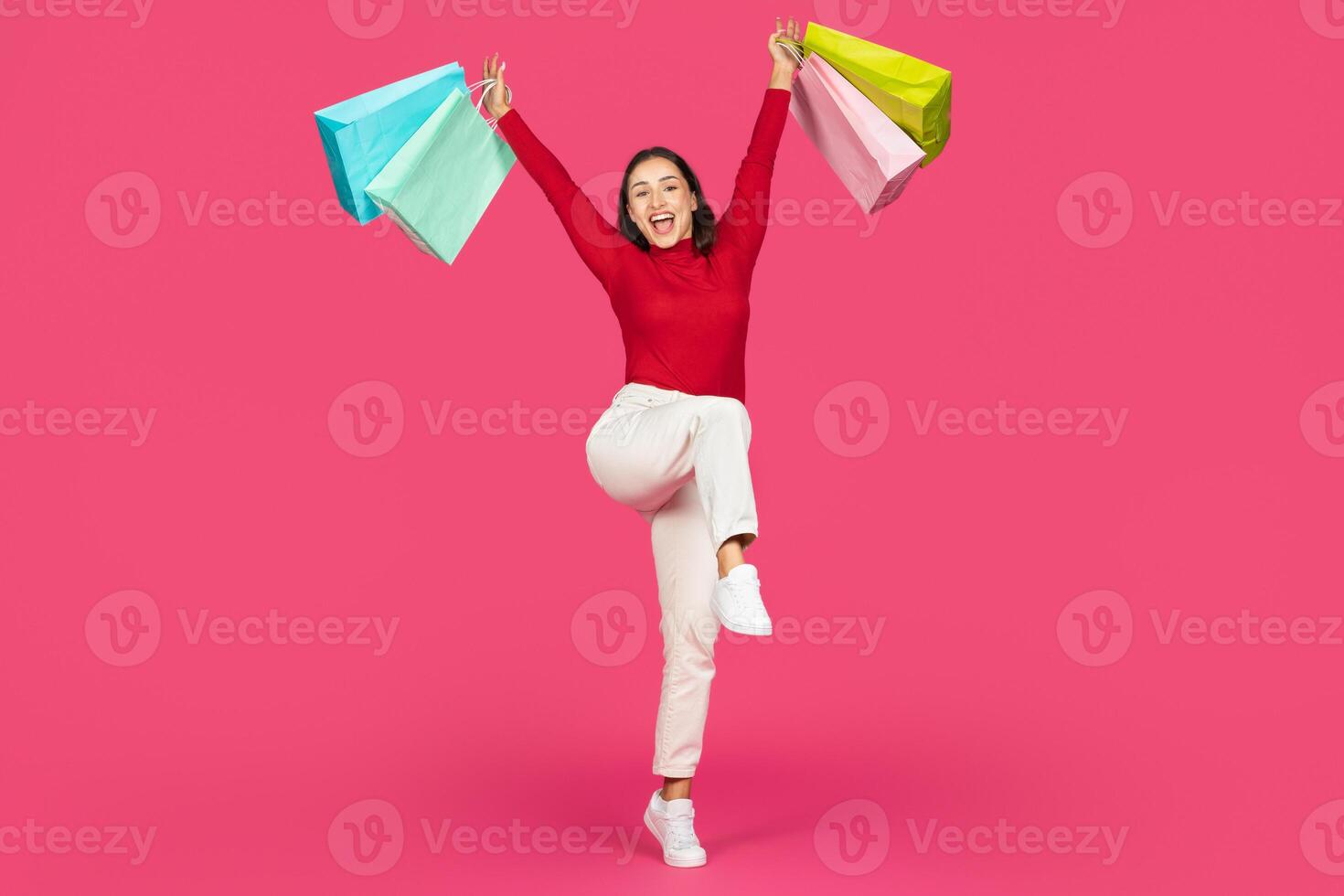 A young woman in a red sweater and white pants jumps happily with colorful shopping bags in each hand, set against a vibrant pink background. Her expression radiates excitement and joy. photo