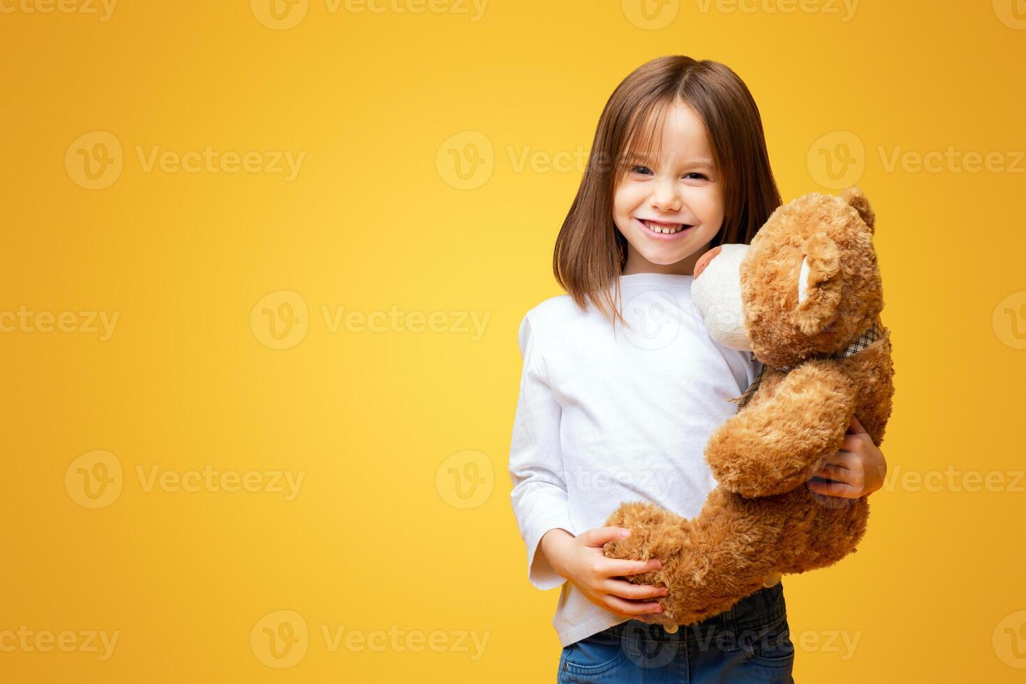 Smiling beautiful little girl embracing big teddy bear over yellow background, kids toys concept photo