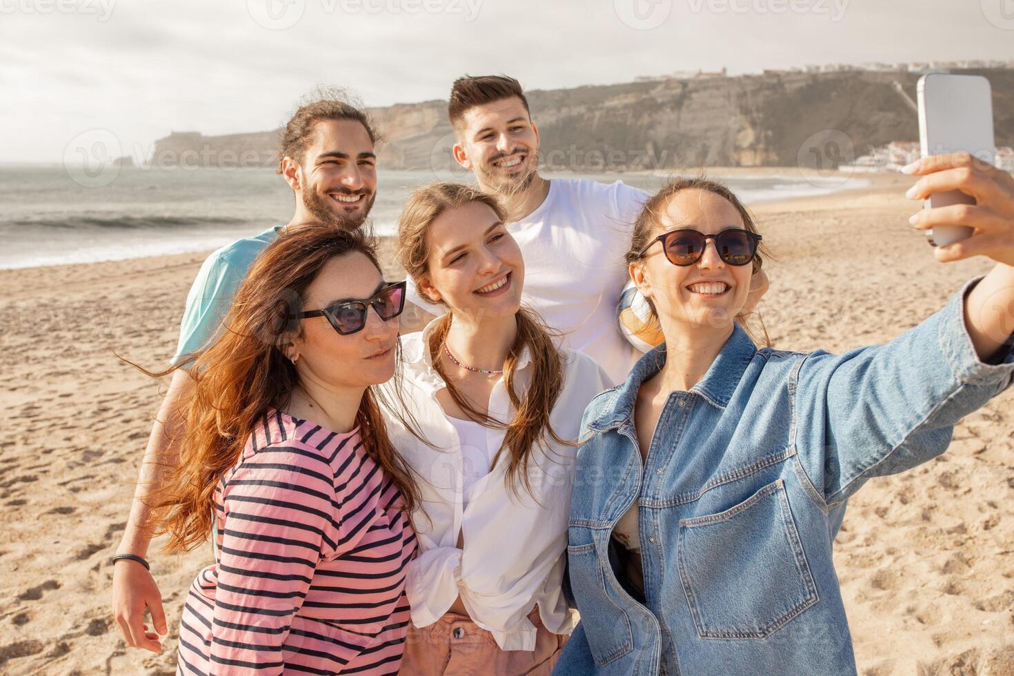 A group of five friends smiles happily as they take a selfie on a sandy beach. The sun sets in the background, casting golden light over the cheerful gathering. photo