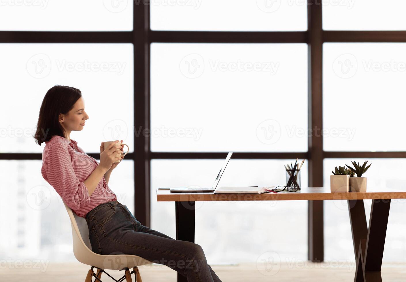 Coworking Concept. Side view of young female manager in shirt sitting at table, drinking coffee and working on laptop photo