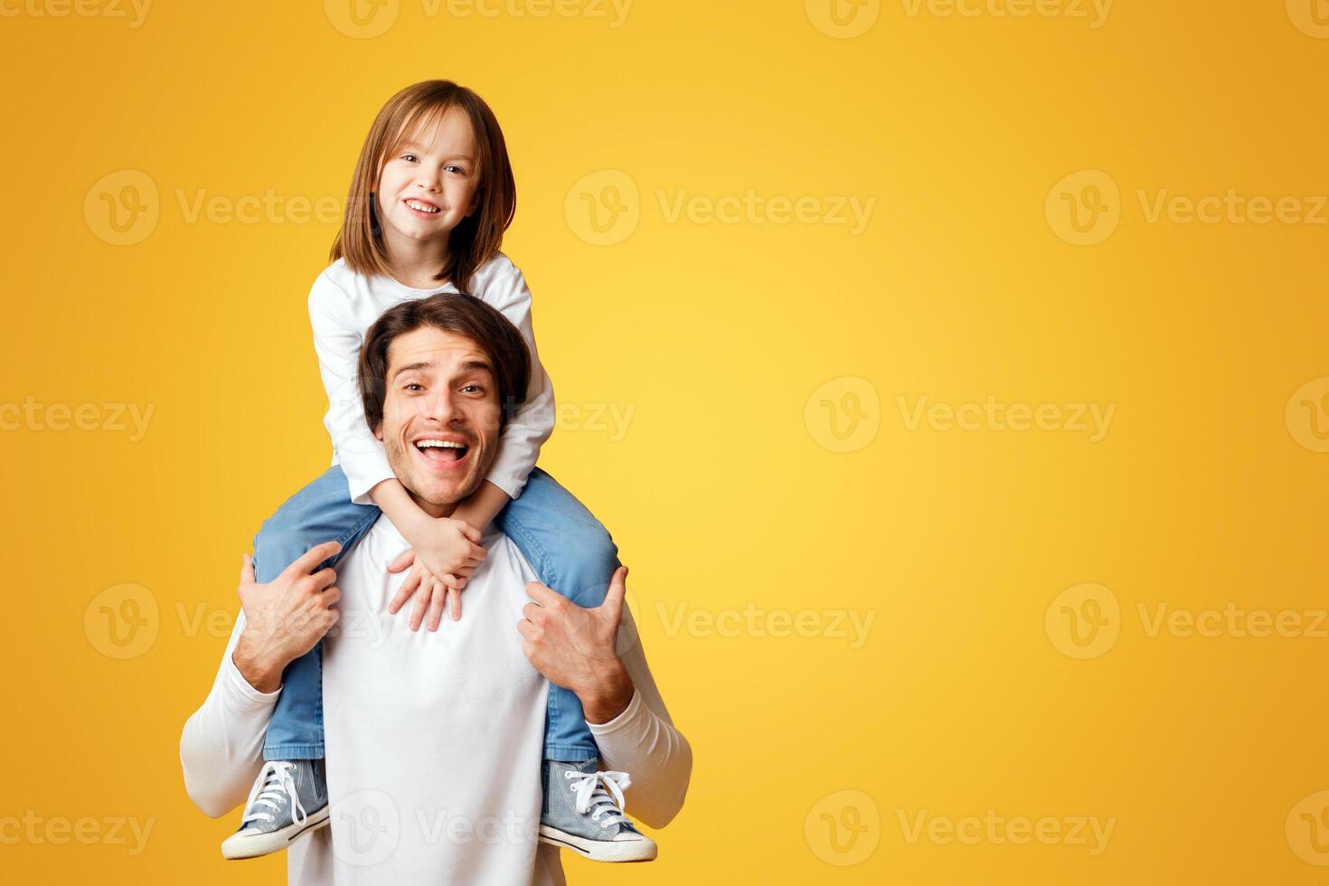 Handsome dad carrying his lovely cheerful little daughter on shoulders, posing over yellow background, copy space photo