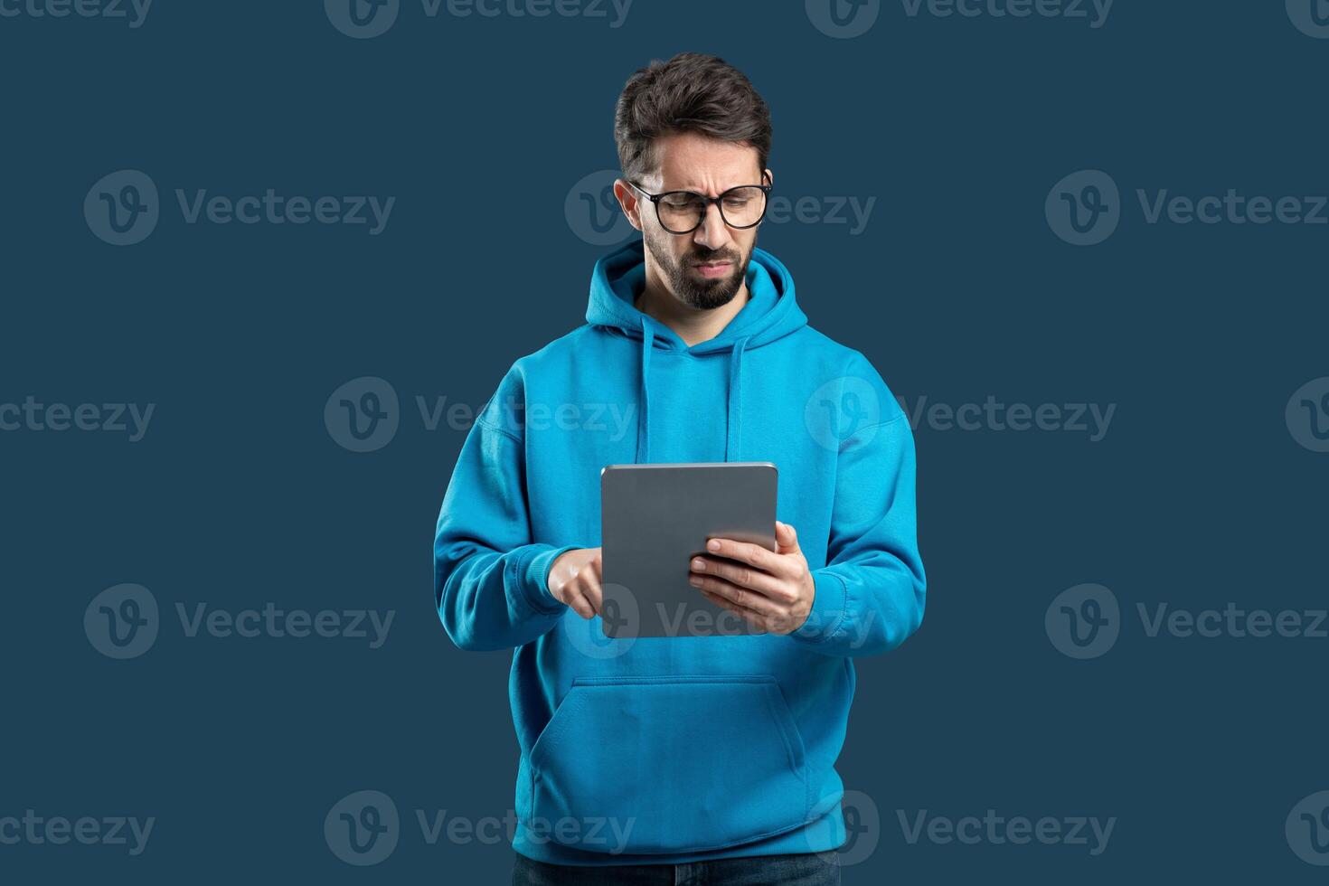 A young man wearing a blue hoodie focuses on a tablet with a serious expression, standing against a solid dark background. He appears to be engaged in thoughtful activity with the device. photo