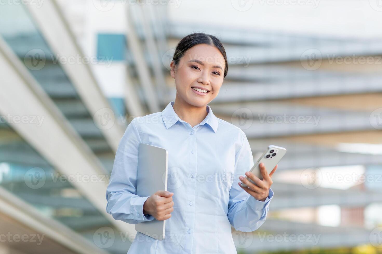 A young Asian woman professional stands outside a contemporary building, smiling while holding a smartphone in one hand and a laptop in the other, showcasing a confident and productive demeanor. photo