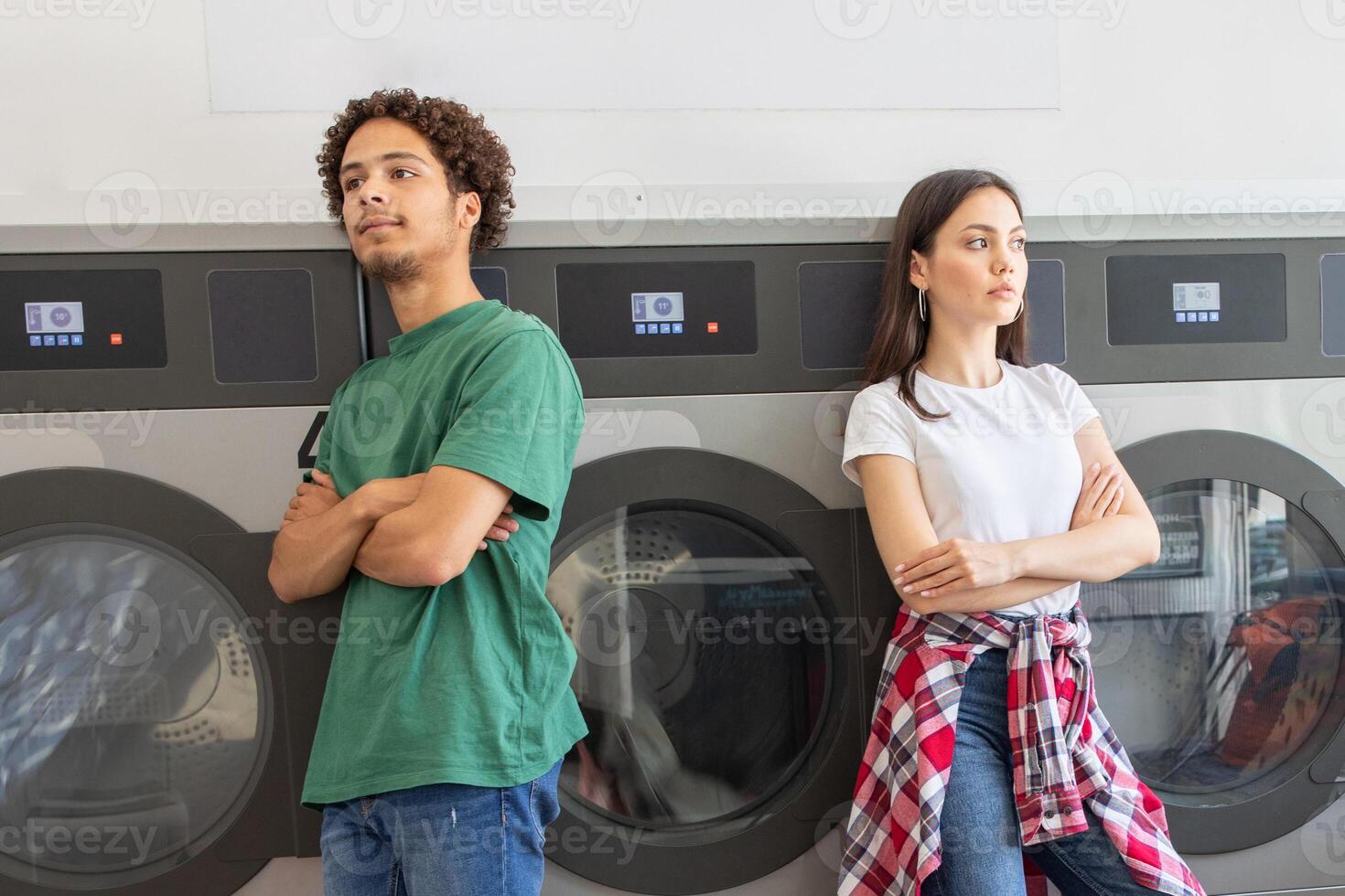 A couple waits at a laundromat, standing on opposite sides while their clothes wash. The atmosphere is tense, reflecting their current emotional distance amid a mundane chore. photo