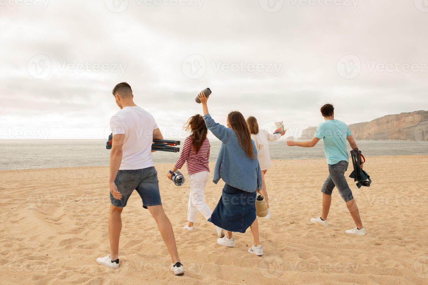 A group of friends walks along the sandy beach, holding cameras and other items, enjoying a fun outing during sunset. The ocean waves gently lap against the shore in the background. photo