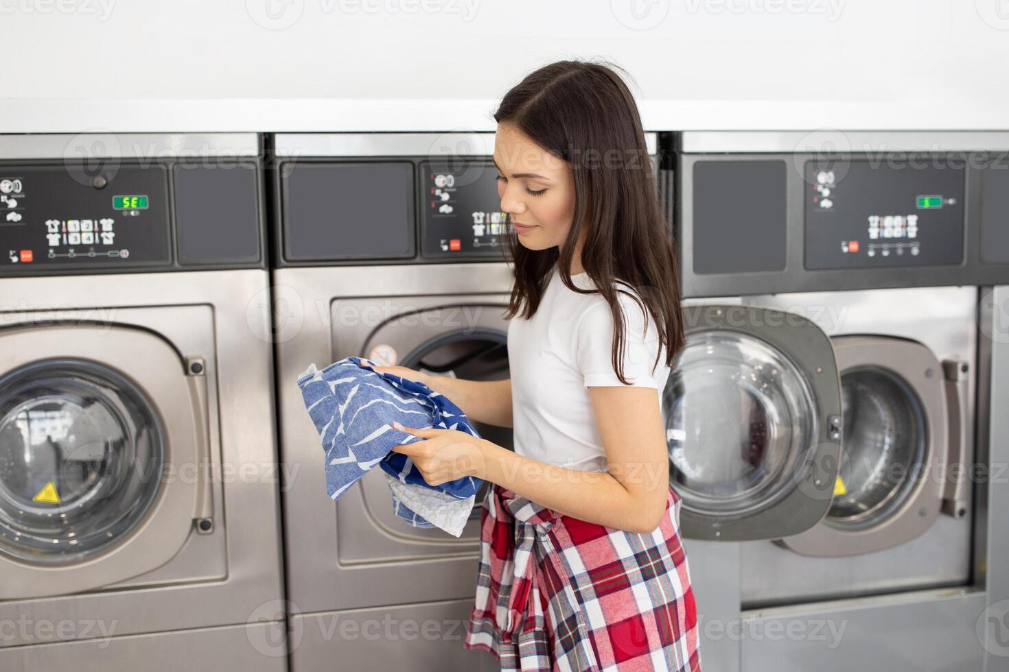 A young woman stands in a self-service laundromat, carefully sorting through clothes. She holds a neatly folded blue item while nearby washing machines await her next move. photo