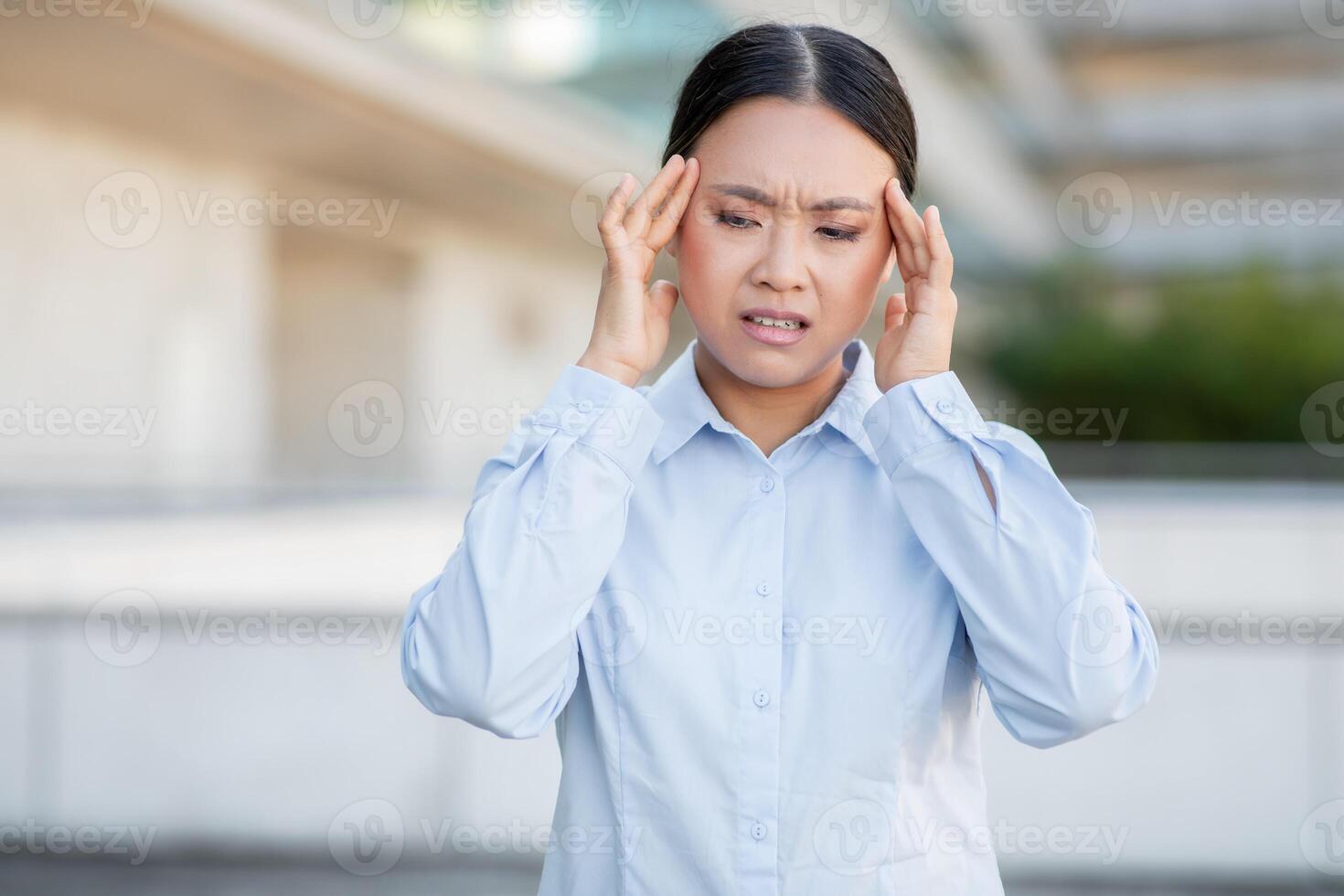 A young Asian woman is standing outside in an urban environment, holding her head with a look of discomfort, likely dealing with a headache or stress, while dressed in a professional outfit. photo
