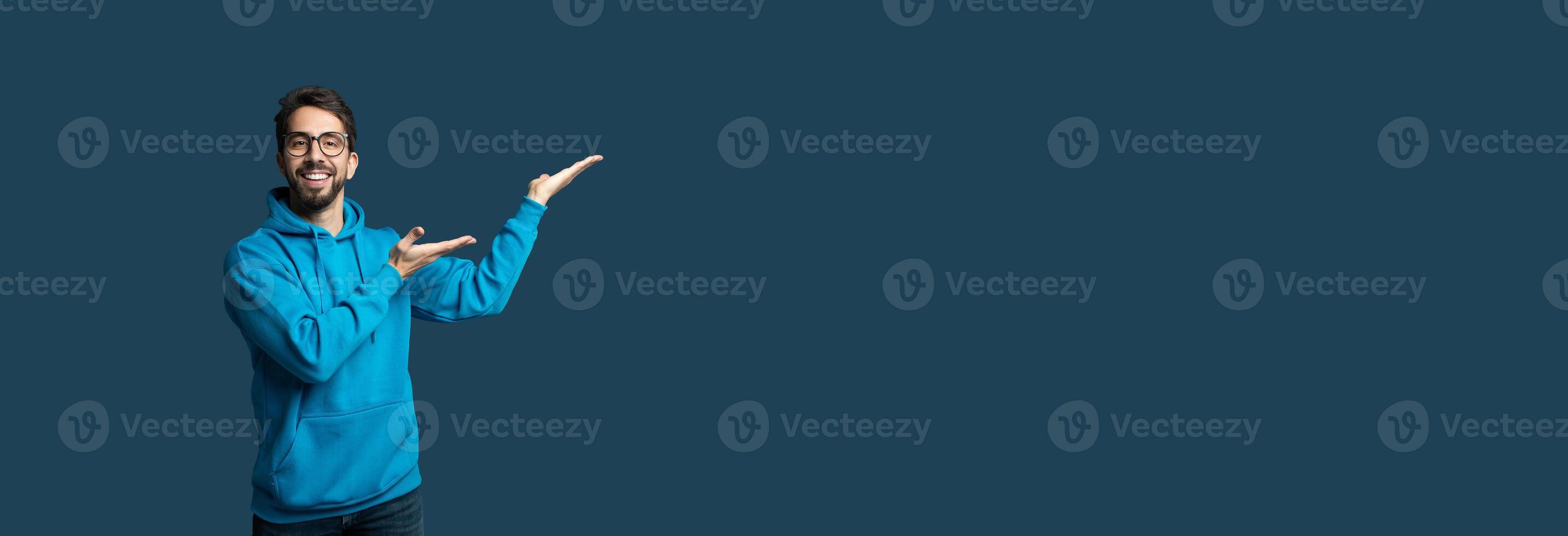 A man wearing a vibrant blue hoodie smiles and gestures with his hands, inviting viewer engagement against a solid blue background, panorama with copy space photo