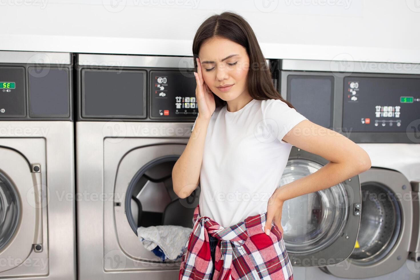 A woman stands in a laundromat, visibly frustrated and holding her head with one hand, while the washing machines hum in the background during the afternoon. photo