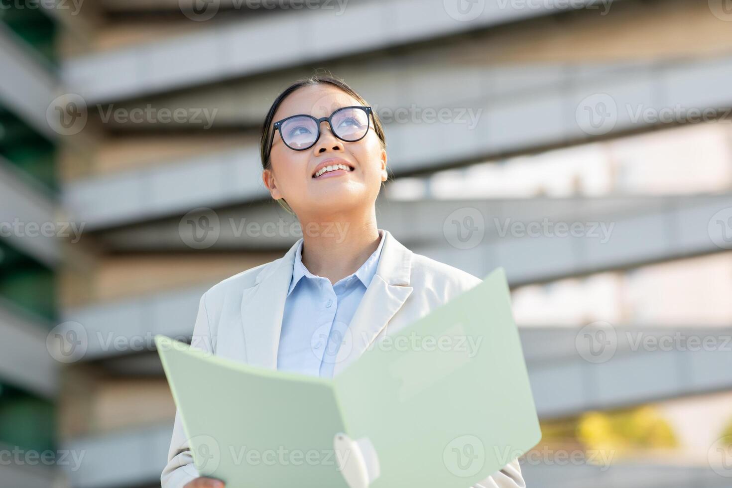 Asian woman dressed in business attire looks up with a smile while holding a folder outside a contemporary building. She appears confident and engaged, enjoying her surroundings. photo
