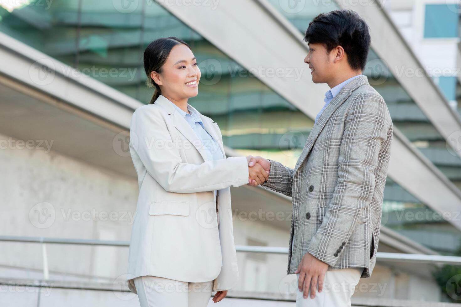 Two Asian business professionals in formal attire shake hands outside a contemporary office building, showcasing a moment of agreement or partnership during a daytime meeting. photo