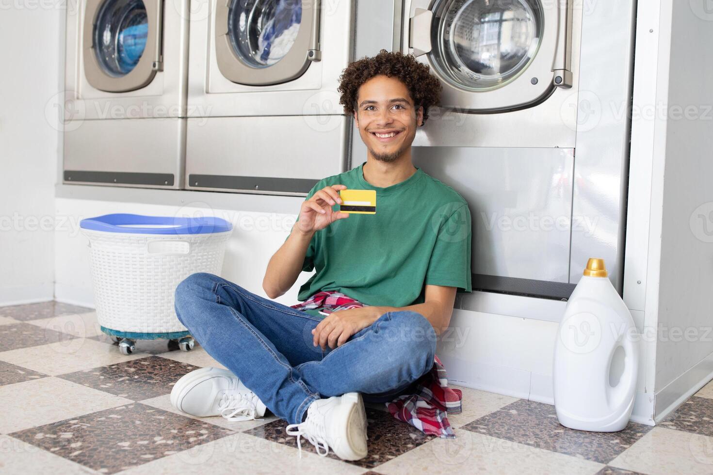 A young man sits on the floor of a laundromat, smiling as he holds a laundry card in his hand. Washing machines are visible in the background, creating a casual atmosphere. photo