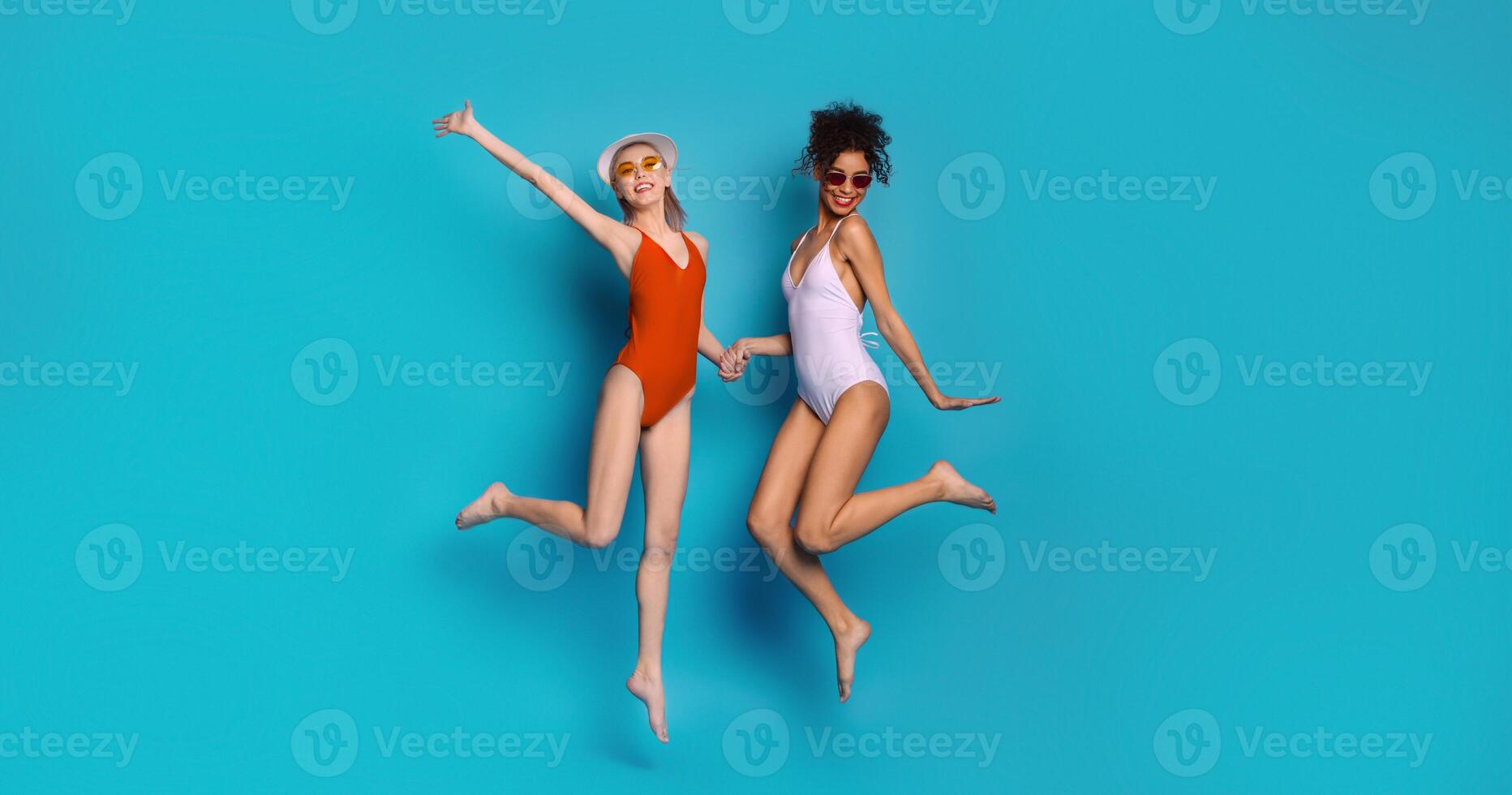 Positive multiethnic ladies in swimsuits jumping on blue background, enjoying summer holidays together photo