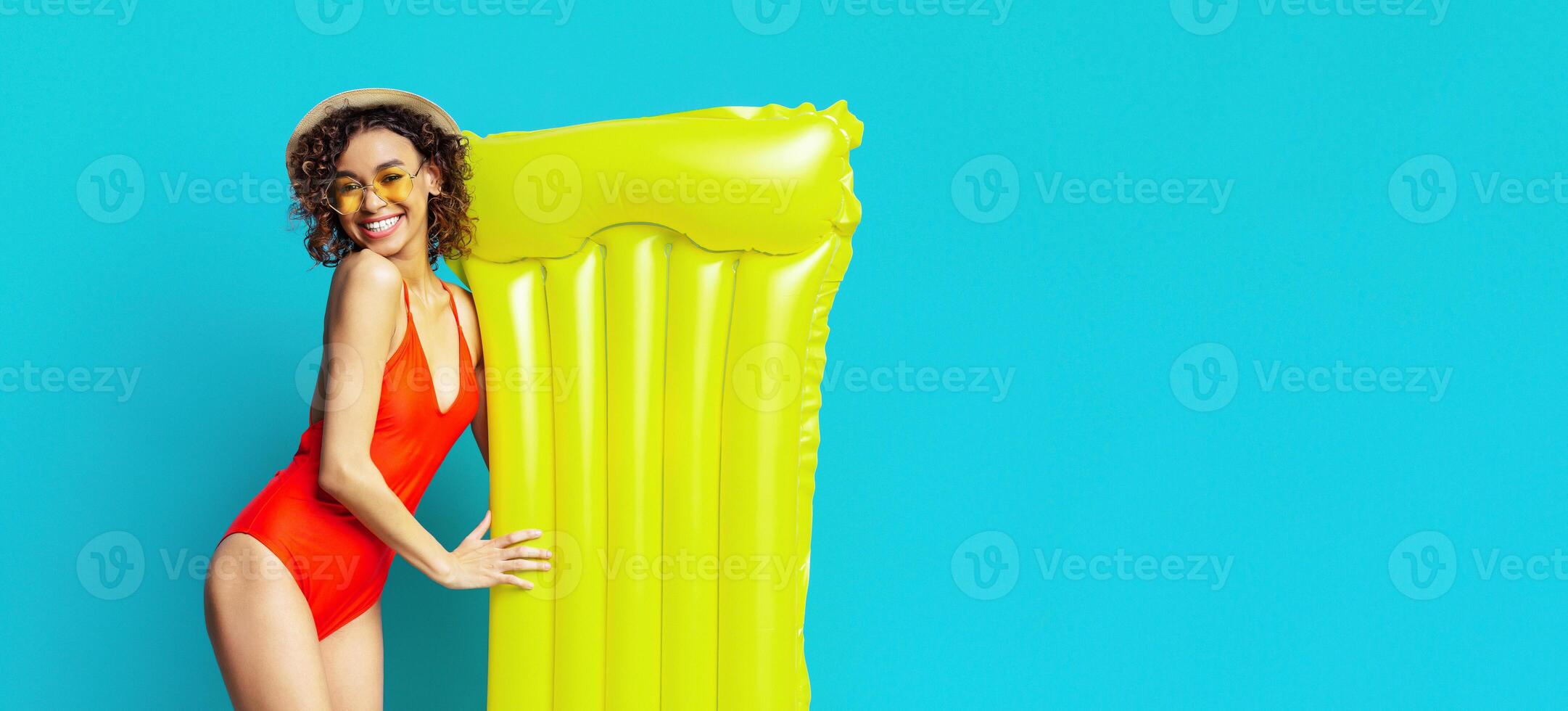 A young black woman in a red swimsuit and straw hat smiles while holding a yellow inflatable raft in front of a bright blue background. She is looking directly at the camera, copy space photo