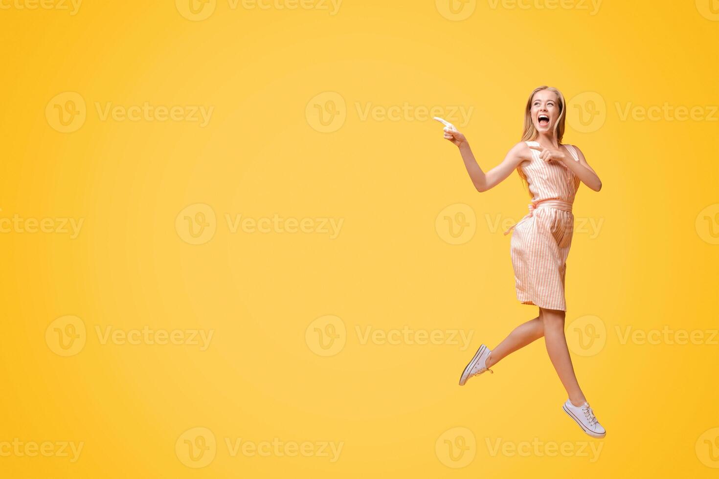 Great Offer. Excited Teen Girl In Summer Dress Jumping In Air And Pointing Aside At Copy Space On Yellow Background photo