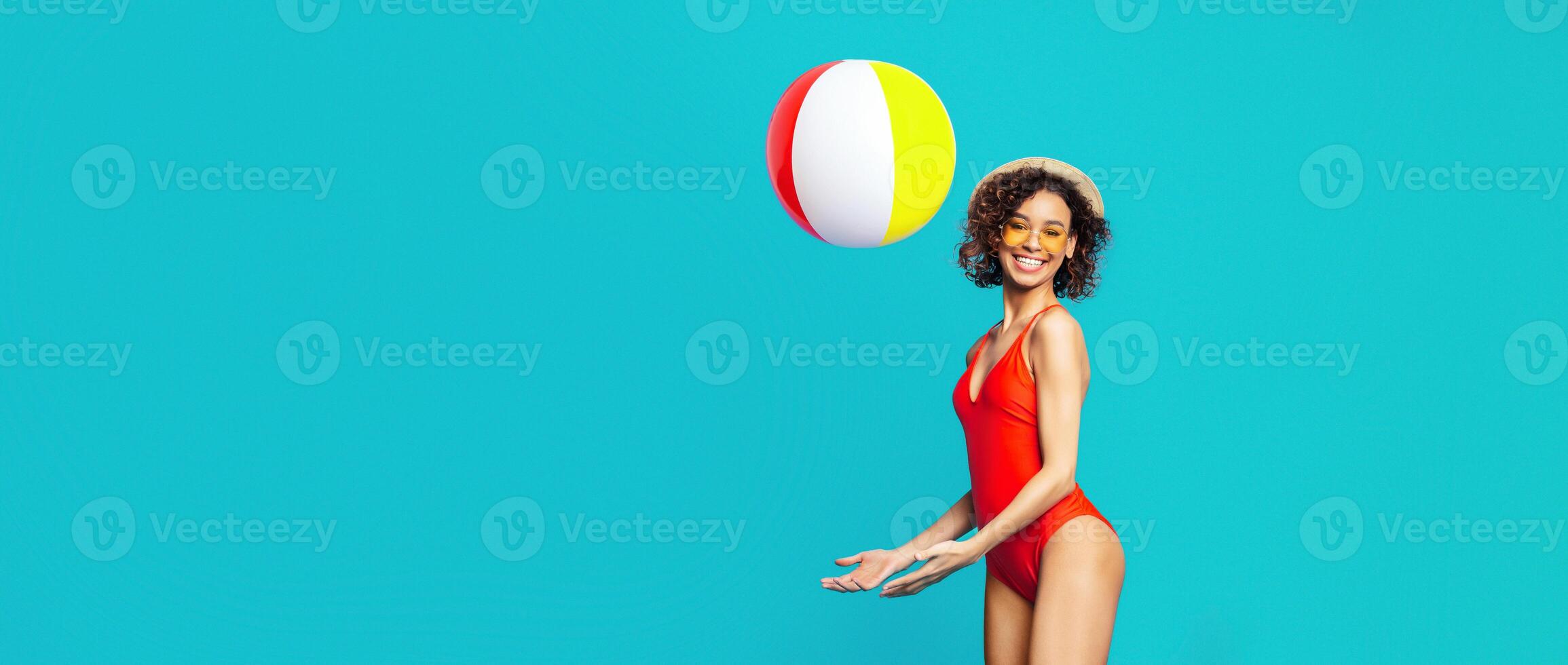 African American young woman in a red swimsuit and a straw hat smiles playfully as she catches a beach ball in a studio with a blue backdrop. The beach ball is red, yellow, and white. photo