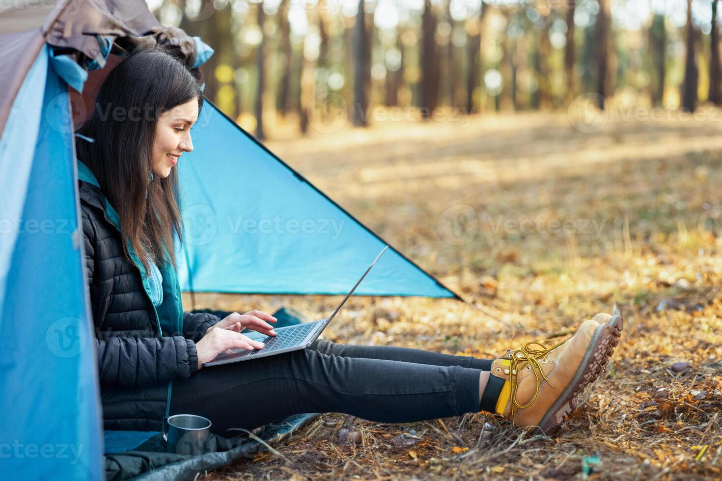 Side view of cheerful girl sitting in tent alone and using laptop, copy space photo