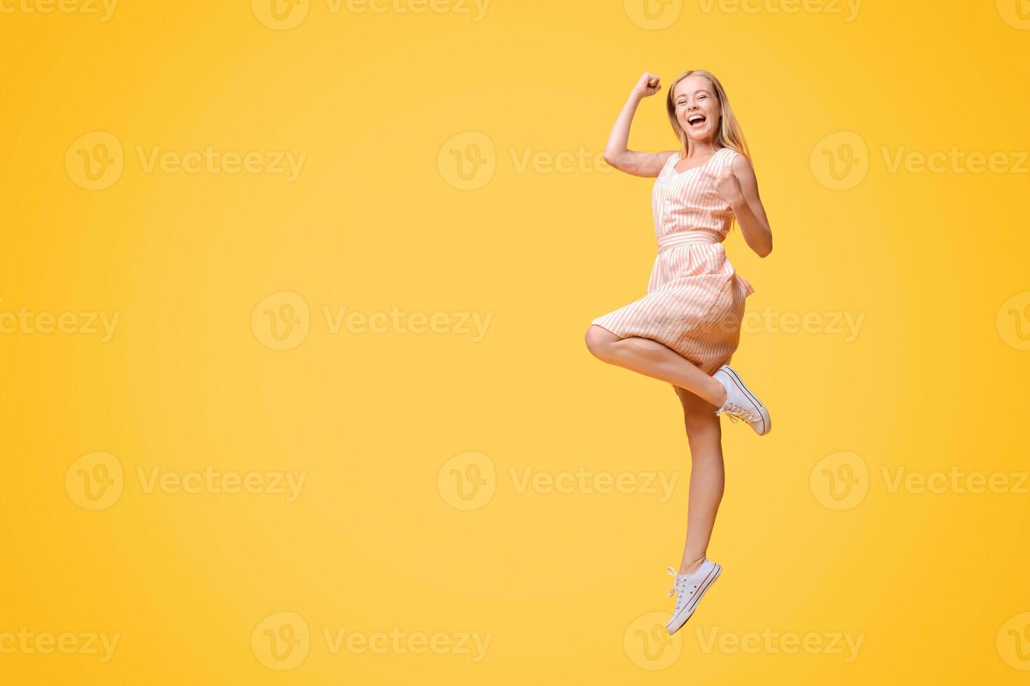 Overjoyed young woman jumping in the air, celebrating success with raised fists over yellow studio background, full-length shot, free space photo